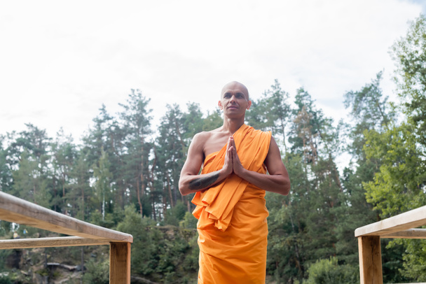 low angle view of buddhist monk meditating with praying hands near wooden fence in forest - Foto, immagini
