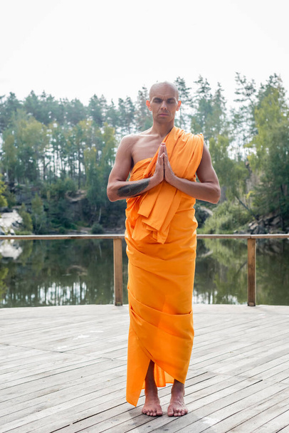 volledige weergave van boeddhistische monnik mediteren met biddende handen op houten platform in de buurt van bos meer - Foto, afbeelding