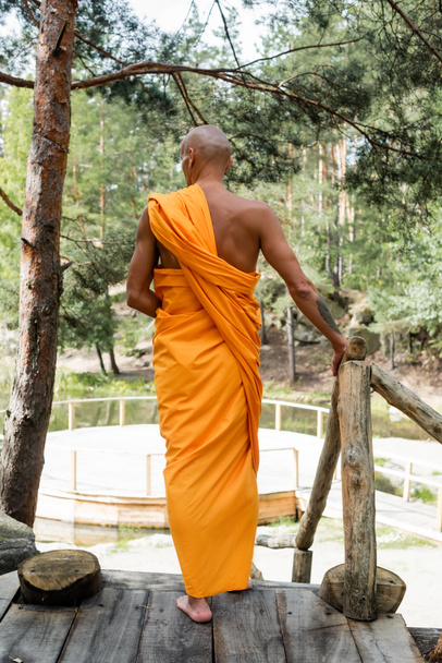 森の中の木製柵の近くに立つ伝統的なオレンジの衣姿の仏教徒の後ろ姿 - 写真・画像