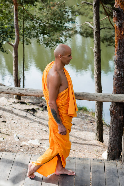 вид збоку буддійського монаха, що йде дерев'яною прогулянкою біля лісового озера
 - Фото, зображення