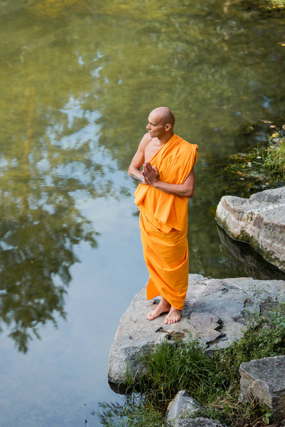 vysoký úhel pohledu bosého buddhistického mnicha v oranžovém rouchu modlícího se poblíž řeky - Fotografie, Obrázek