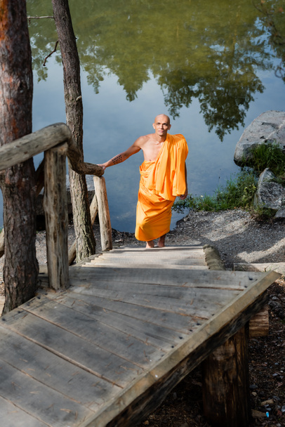 Turuncu cüppeli Budist göl kenarındaki ahşap merdivenlerde yürüyor. - Fotoğraf, Görsel