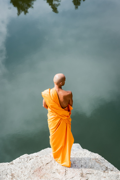 high angle view of buddhist in orange kasaya meditating on rocky cliff over lake  - Photo, Image