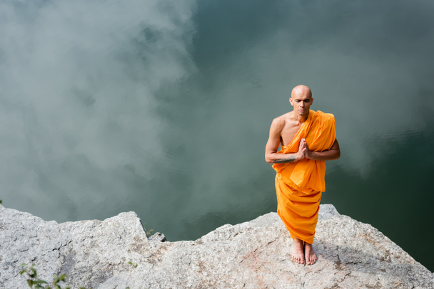 vista de alto ângulo de budista em robe laranja orando em penhasco rochoso sobre a água - Foto, Imagem