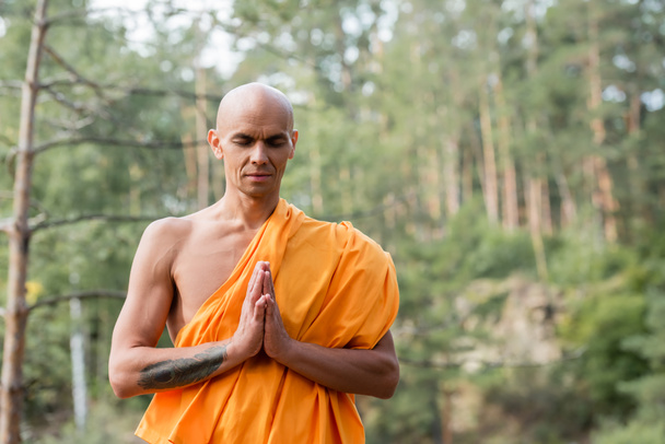 uomo senza peli in vestaglia buddista pregando con gli occhi chiusi nella foresta - Foto, immagini