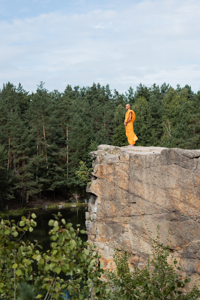 oldalnézetben buddhista narancssárga kasaya meditál magas sziklás sziklafal felett tó erdőben - Fotó, kép