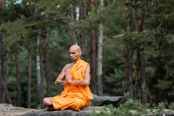buddhist monk in orange kasaya sitting in lotus pose with praying hands while meditating in forest - Фото, изображение