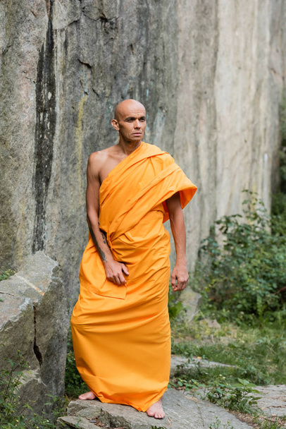 άνδρας με παραδοσιακά βουδιστικά ρούχα κοιτάζει αλλού ενώ στέκεται κοντά στο βράχο - Φωτογραφία, εικόνα