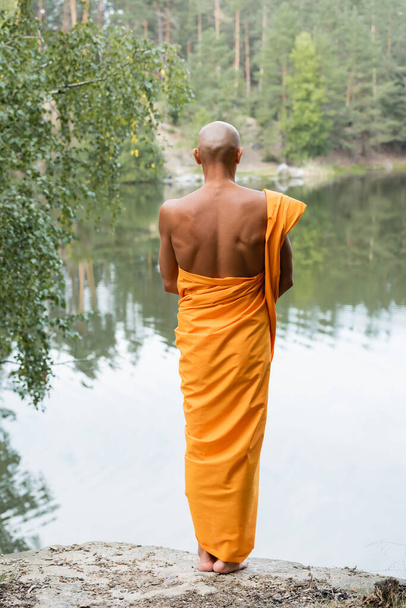 back view of bald man in traditional buddhist robe meditating near forest pond - Фото, изображение
