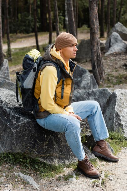 traveler with backpack, wearing warm vest and beanie, sitting on stone in forest - Fotó, kép