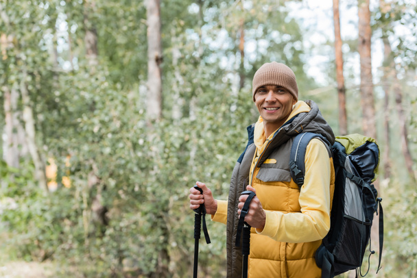 cheerful tourist with backpack and trekking poles looking away in forest - Photo, Image