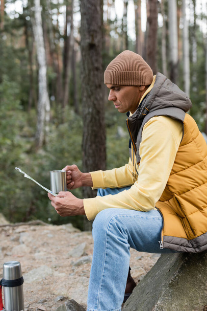 voyageur en bonnet et gilet chaud assis avec tasse en métal et carte en forêt - Photo, image
