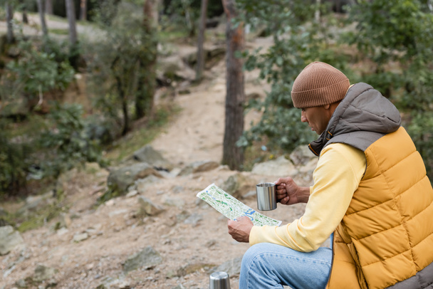 túrázó sapkában és meleg mellény ül az erdőben térképpel és fém bögre - Fotó, kép