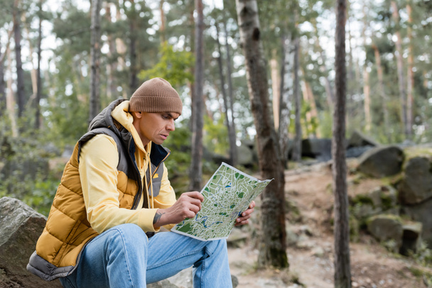 túrázó őszi ruhában ül a kövön az erdőben, és nézi a térképet - Fotó, kép