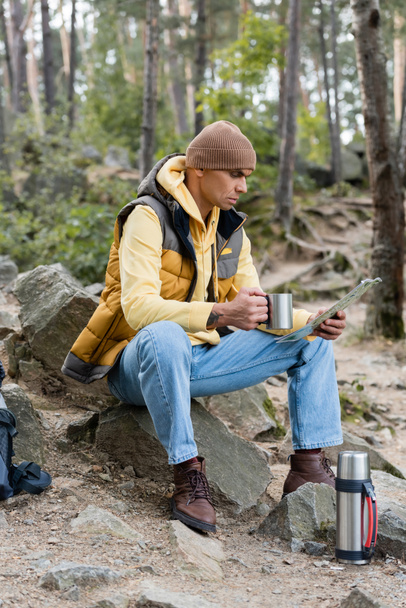 турист смотрит на карту, сидя с металлической кружкой в лесу - Фото, изображение