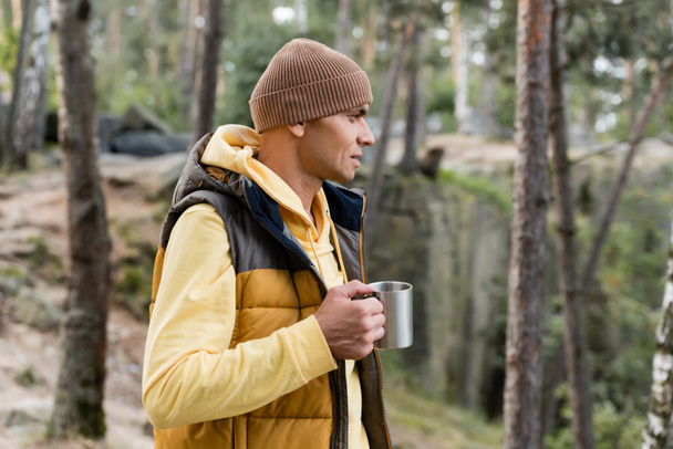 tourist in warm vest and beanie holding metal mug in forest - Zdjęcie, obraz