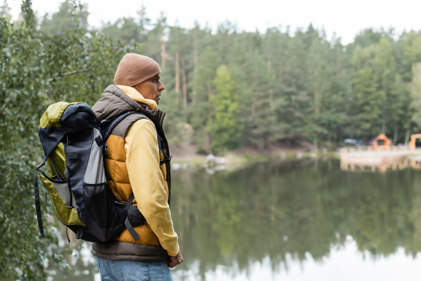 hiker with backpack looking away while standing near lake in forest - 写真・画像