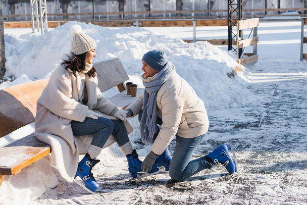 happy man wearing ice skates on cheerful girlfriend in winter hat - Photo, Image
