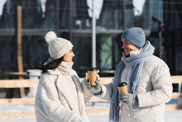 Счастливый молодой человек дает бумажную чашку веселой девушке в зимней шляпе - Фото, изображение