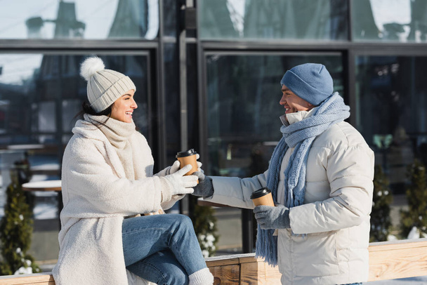 joyful young man giving paper cup to cheerful girlfriend in winter hat - Photo, image