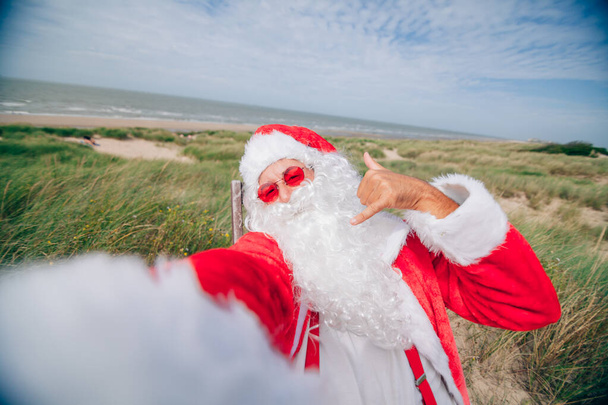 Santa Claus en el mar haciendo un vídeo con su teléfono móvil mientras hace el gesto de colgar suelto con la mano - Foto, Imagen