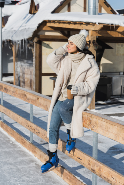 longitud completa de la joven feliz en sombrero de invierno y patines de hielo sosteniendo taza de papel cerca de pista de hielo - Foto, Imagen