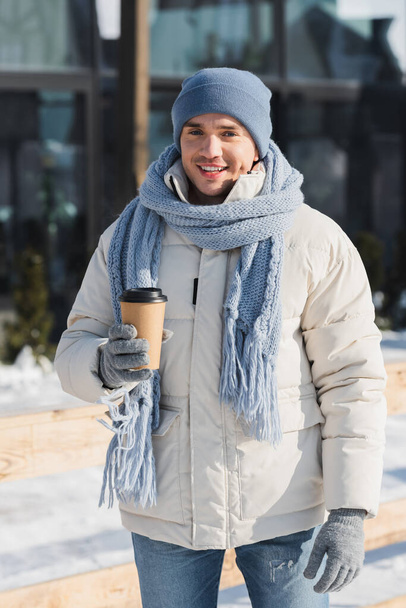 Kışlık şapkalı ve eşarplı mutlu genç adam elinde kağıt bardakla dışarıda - Fotoğraf, Görsel