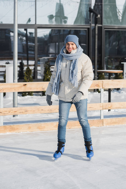 full length of happy young man in scarf, winter hat and ice skates on ice rink - Fotoğraf, Görsel