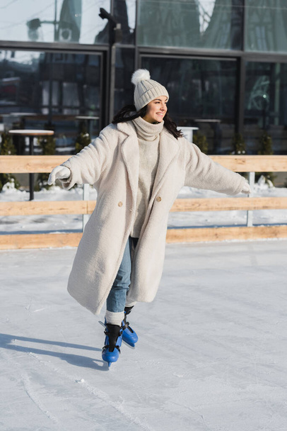 longitud completa de la joven feliz en sombrero de invierno y capa de patinaje en pista de hielo - Foto, Imagen