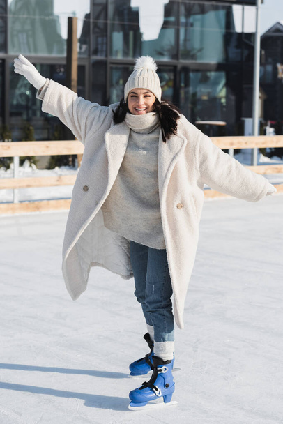 full length of pleased young woman in winter hat and coat skating on ice rink - 写真・画像
