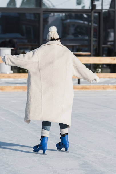 back view of young woman in winter hat and coat skating on ice rink - Фото, изображение