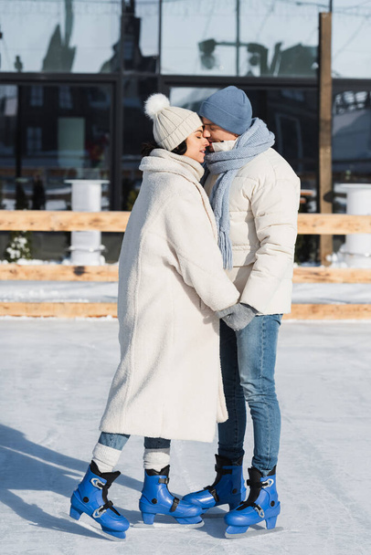 full length of young couple in winter hats holding hands while kissing on ice rink - 写真・画像