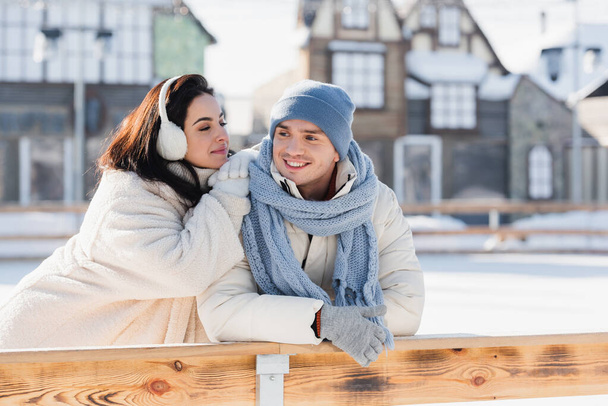 young woman in ear muffs and cheerful man in winter hat leaning on wooden border on ice rink  - Fotografie, Obrázek