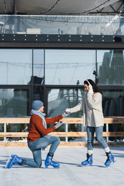 volledige lengte van de man knielen en houden doos met ring, terwijl het maken van aanzoek aan gelukkige vrouw in oor moffen op de ijsbaan - Foto, afbeelding