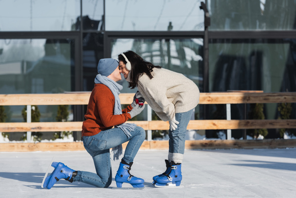 full length of young man holding box with wedding ring and kneeling while making proposal and kissing with woman on ice rink - Fotó, kép