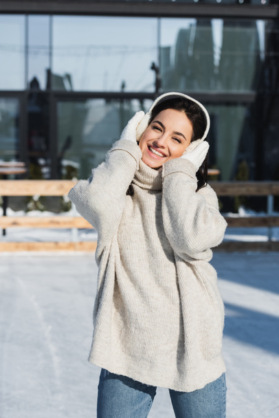 alegre joven mujer ajustando orejeras en pista de hielo - Foto, Imagen