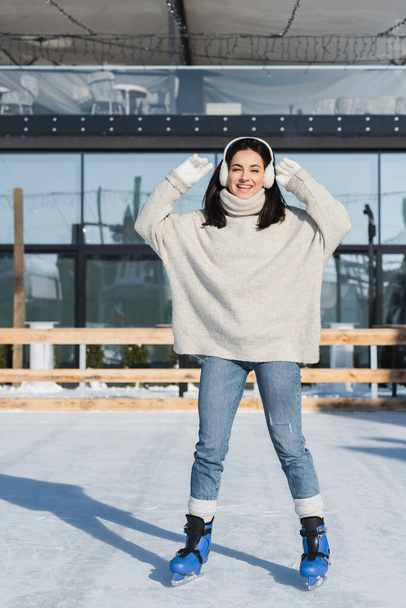 πλήρες μήκος χαρούμενη νεαρή γυναίκα σε πουλόβερ και ωτοασπίδες πατινάζ σε παγοδρόμιο - Φωτογραφία, εικόνα