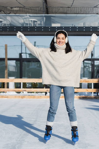 full length of happy young woman in sweater and ear muffs skating on ice rink - Foto, Bild