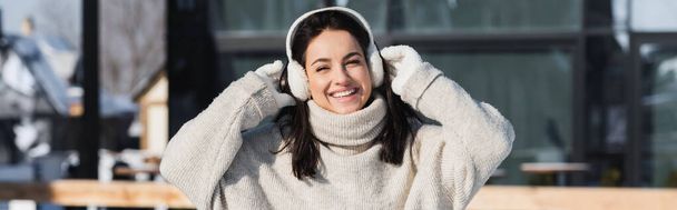 vrolijke jonge vrouw in trui en oorkappen in de winter, banner - Foto, afbeelding