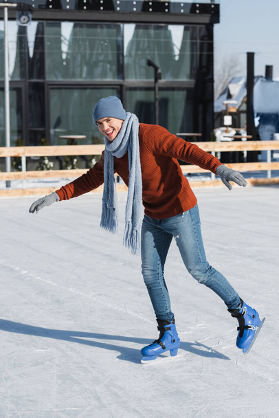 longitud completa de hombre joven feliz en suéter, bufanda y sombrero de invierno patinaje en pista de hielo - Foto, Imagen