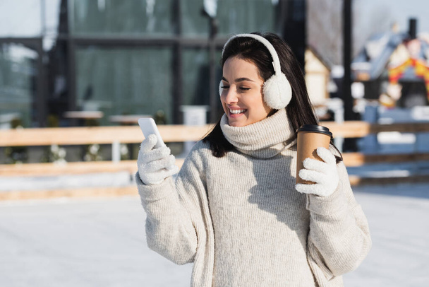 happy young woman in sweater and ear muffs holding paper cup and using smartphone on ice rink  - Foto, Imagen