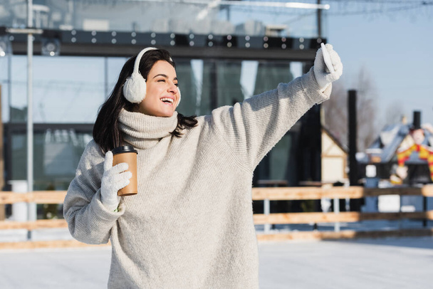 mulher feliz em suéter e abafadores de ouvido segurando copo de papel e tomando selfie na pista de gelo  - Foto, Imagem