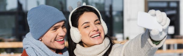 happy woman in sweater and ear muffs taking selfie with boyfriend in wintertime, banner - Photo, Image