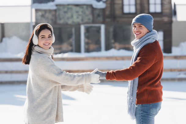 sonriente joven mujer en orejeras cogido de la mano con alegre novio en pista de hielo  - Foto, Imagen
