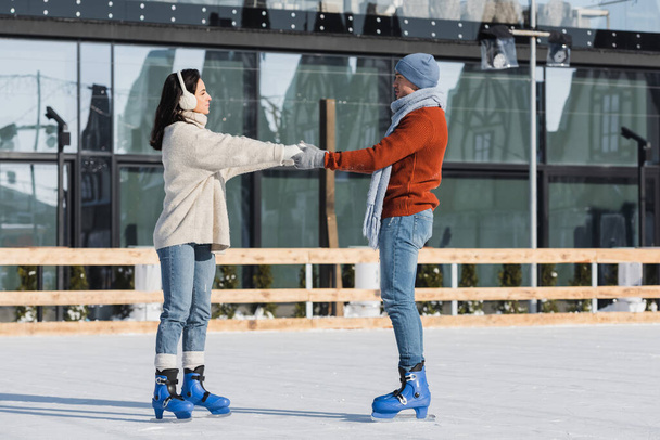 full length of young woman in ear muffs holding hands with boyfriend on ice rink - Foto, imagen