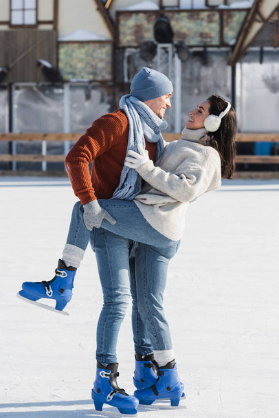 longitud completa de hombre alegre levantando pierna de novia feliz en pista de hielo  - Foto, Imagen