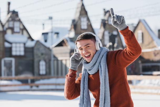 happy young man in sweater and scarf listening music while singing and pointing with finger  - 写真・画像