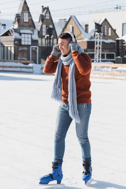 full length of cheerful young man in sweater and scarf listening music while skating on ice rink  - Foto, Imagem