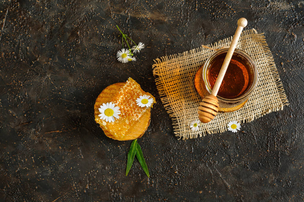 Honeycomb and jar of honey with wooden spoon top view, dark background, flowers and copy space - Photo, image