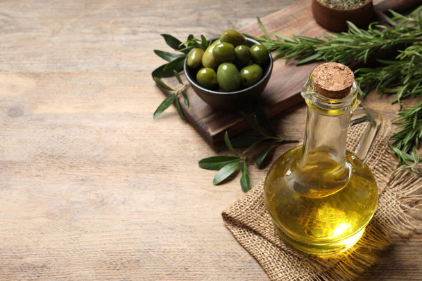 Glass jug of oil, ripe olives and green leaves on wooden table. Space for text - Foto, Bild
